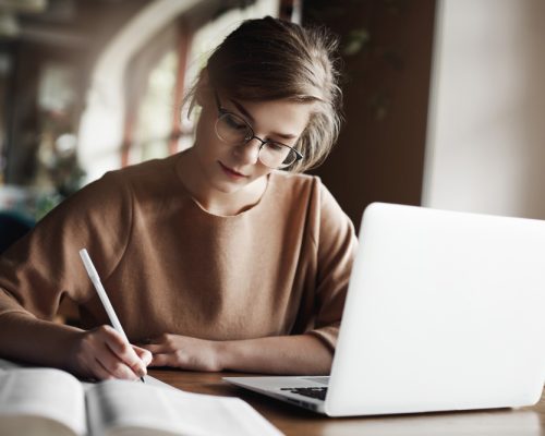 hardworking-focused-woman-trendy-glasses-concentrating-writing-essay-sitting-cozy-cafe-near-laptop-working-making-notes-carefully